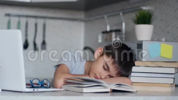 疲惫的小学生坐在笔记本电脑和课本旁边的书桌前做作业睡着了视频的预览图