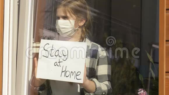 戴着医用面具的女孩站在窗口在家里贴着一张海报检疫视频的预览图