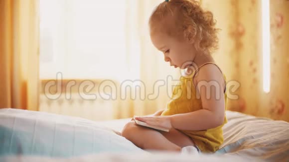 专注的女婴在家里阳光明媚的房间里看着小书里的照片视频的预览图