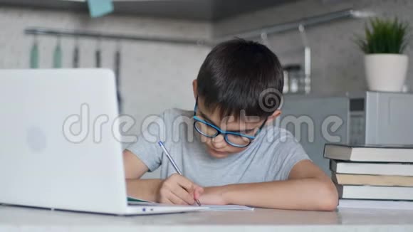 戴眼镜的孩子在家里坐在餐桌旁的厨房里用笔记本电脑书和笔记本上课视频的预览图