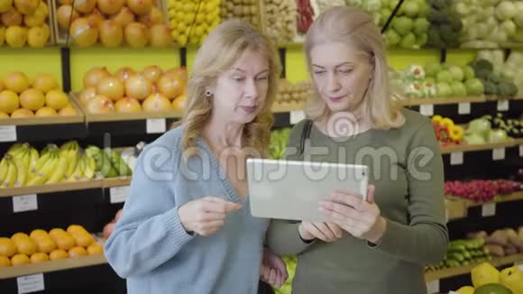 积极的高加索老年妇女在杂货店使用平板电脑的肖像两个家庭主妇站在商品和视频的预览图
