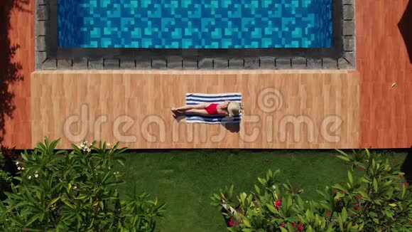 空中美丽的年轻女子在游泳池附近的毛巾上日光浴穿着红色比基尼戴着热带草帽在那里玩耍视频的预览图