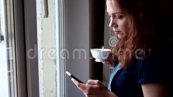 年轻女子手里拿着电话喝咖啡在隔离期间看着窗外社会距离视频的预览图