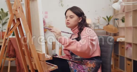 十几岁的孩子穿着艺术家的围裙在她的房间里一只手拿着画笔患有唐氏综合症的女孩视频的预览图
