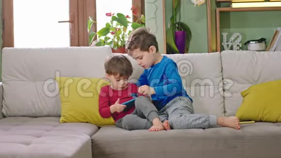 两个可爱的孩子坐在沙发上拿着智能手机一起玩游戏度过快乐的时光视频的预览图