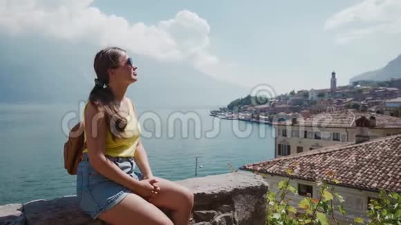 戴墨镜的可爱旅游女孩坐在石阶上欣赏小镇景色视频的预览图