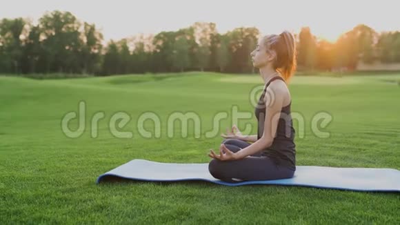少女坐在绿草如茵的莲花位置瑜伽课在夕阳的时候视频的预览图