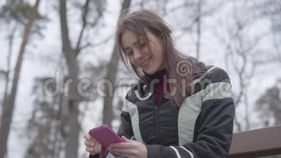 快乐美丽的女孩使用智能手机和微笑坐在公园的长凳上的肖像年轻的黑发女人享受视频的预览图