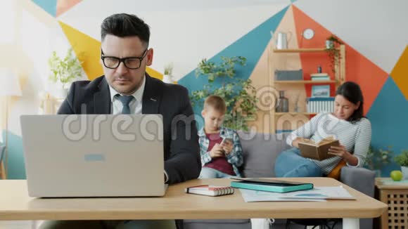 严肃的商人在家里使用笔记本电脑工作妻子和孩子在背景中可以看到视频的预览图