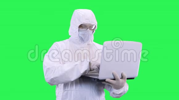 一个戴着面具和眼镜的男人戴着绿色背景的防护服环顾四周将数据输入笔记本电脑视频的预览图