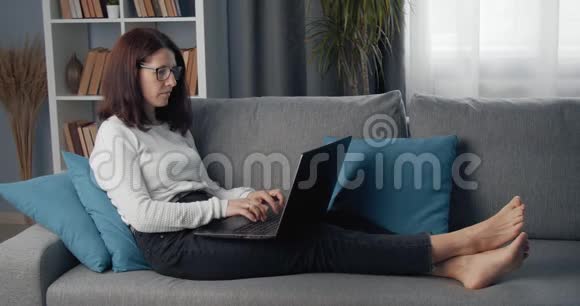 躺在沙发上使用笔记本电脑的成熟女人的侧视图视频的预览图