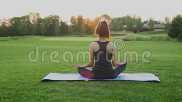 少女坐在绿草如茵的莲花位置瑜伽课在夕阳的时候视频的预览图