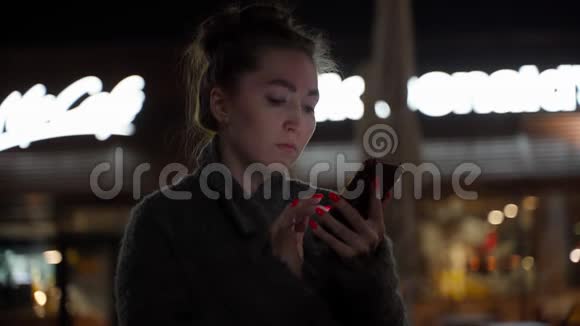 穿着外套手持手机站在夜城大街上的漂亮年轻时髦女人对夜城做出了愉快的反应视频的预览图