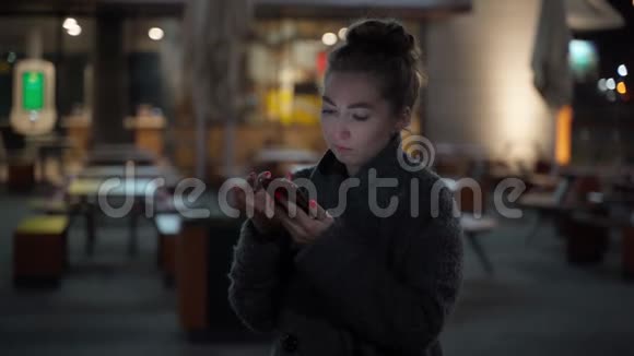 穿着外套手持手机站在夜城大街上的漂亮年轻时髦女人对夜城做出了愉快的反应视频的预览图