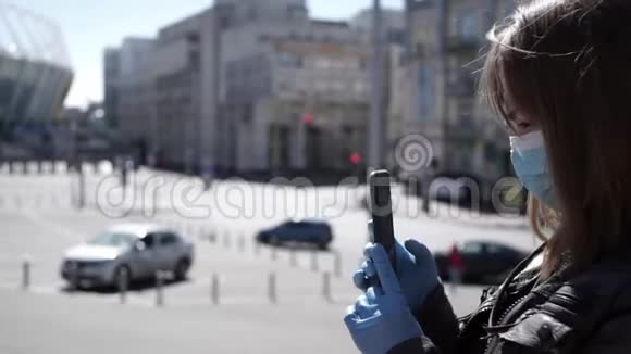 戴着外科口罩和手套的年轻女性在冠状病毒流行期间在户外智能手机上录制视频视频的预览图