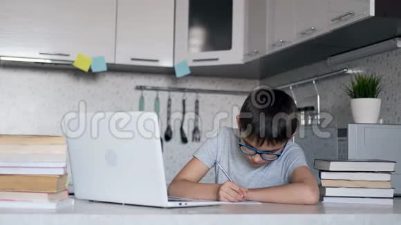 戴眼镜的孩子在家里坐在餐桌旁的厨房里用笔记本电脑书籍和笔记本上课视频的预览图