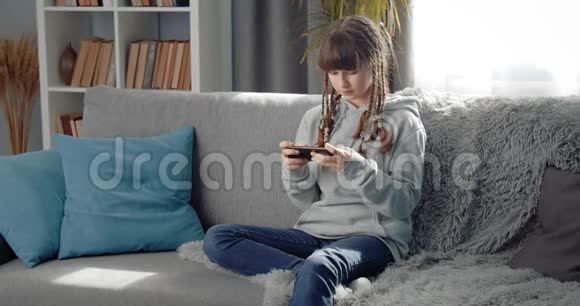 年轻漂亮的女孩在沙发上用智能手机放松视频的预览图