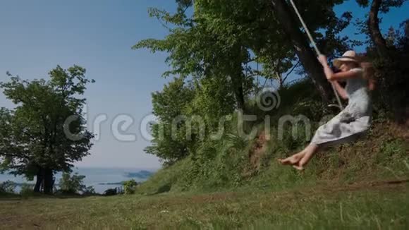 可爱的女人穿着夏装草帽摆动在树秋千侧视长期射击视频的预览图