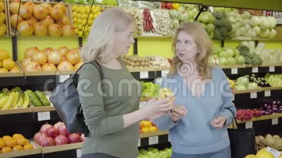 两个成熟的白种人家庭主妇在杂货店里闻着新鲜的黄梨说话自信的女人讨论新鲜事物视频的预览图