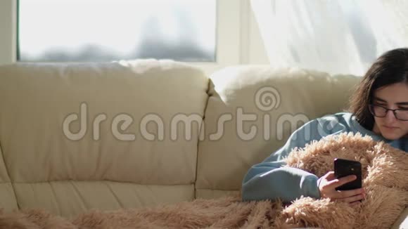 戴眼镜的女孩坐在沙发上拿着手机休息的概念摄像机移动视频的预览图
