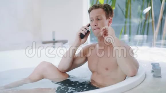 坐在豪华公寓的浴缸里一个肌肉发达的金发男人在谈论智能手机视频的预览图