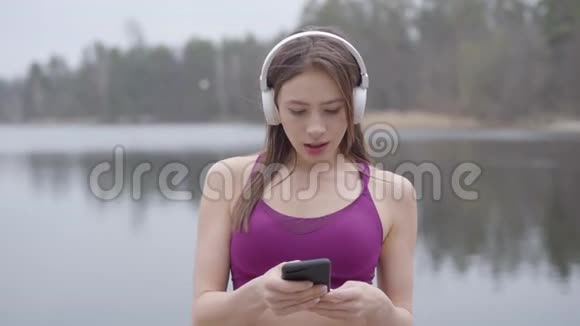 戴着耳机和运动服的可爱年轻女人在湖岸跳舞唱歌的肖像积极的视频的预览图