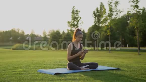 一个年轻的女孩在做瑜伽后坐在绿草上用手机视频的预览图
