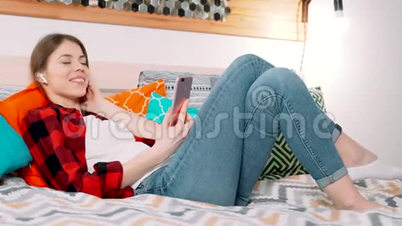 一个年轻迷人的白人女人躺在床上在现代卧室里微笑一边听着用现代耳塞沉思视频的预览图