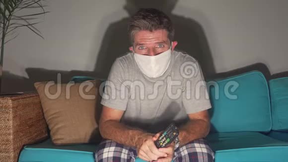 Covid19病毒恐慌锁定悲伤和焦虑的男人戴着医疗面具看电视新闻冠状病毒爆发视频的预览图