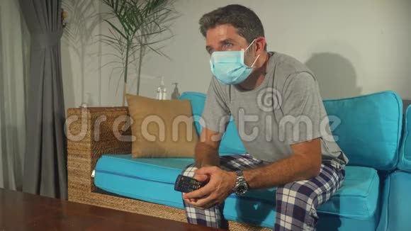 Covid19病毒恐慌锁定悲伤和焦虑的男人戴着医疗面具看电视新闻冠状病毒爆发视频的预览图