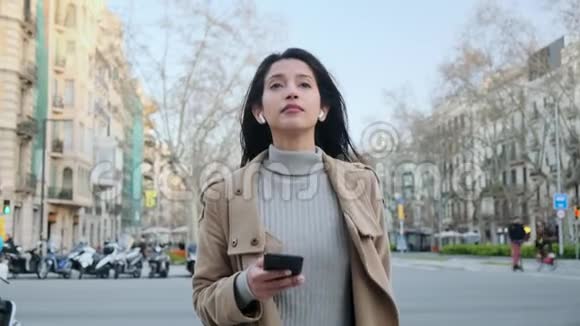 漂亮的年轻女人在街上散步时用手机和无线耳机听音乐视频的预览图