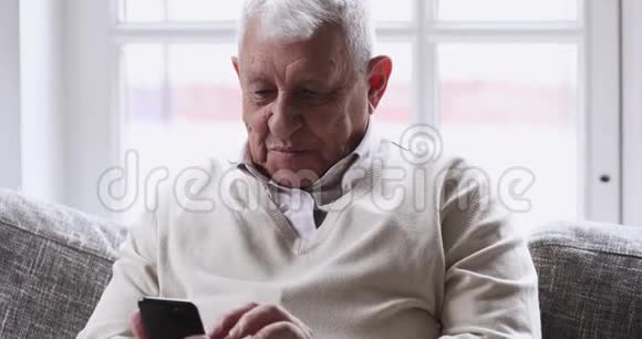 快乐的老人用智能手机在沙发上放松视频的预览图
