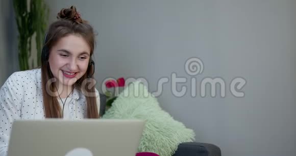 快乐女孩用笔记本电脑和耳机坐在家里客厅的沙发上学习视频的预览图