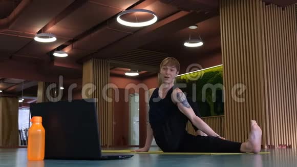 一个身材瘦削手臂肌肉发达的可爱白人在大厅的笔记本电脑前进行在线瑜伽课程广泛视频的预览图