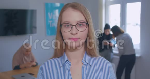 一位年轻漂亮的白种人金发女郎戴着眼镜在办公室背景下大笑的肖像视频的预览图