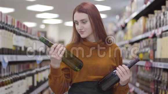 迷人的红发女人看着手中的红酒和白酒把它们拉伸成相机视频的预览图