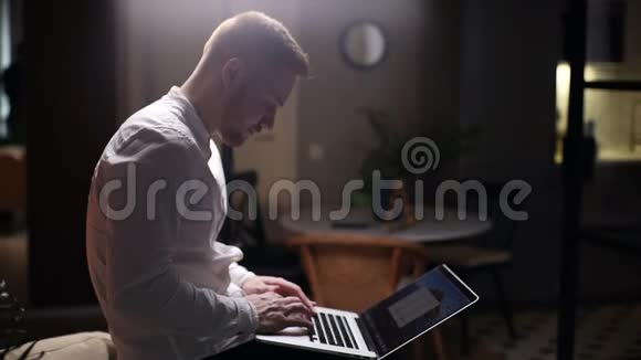 英俊的白种人留着胡须的男人在黑暗的客厅里用笔记本电脑打字跪着视频的预览图