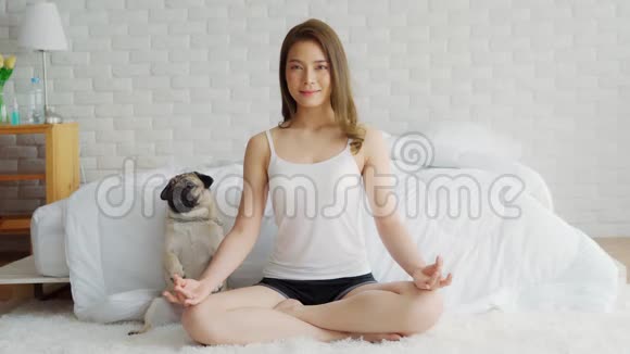 美丽的亚洲女性在家的卧室里练习瑜伽莲花姿势和狗巴格犬享受和放松瑜伽视频的预览图