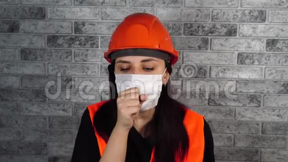 灰色砖墙背景下穿着工作服和医用口罩的女建筑工人咳嗽威胁概念视频的预览图