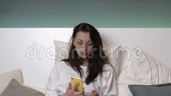 穿着白色睡衣的快乐年轻女子在床上用手机说话微笑着视频的预览图