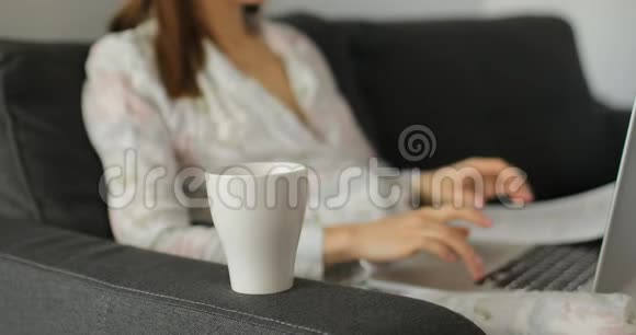 女人在笔记本电脑上工作在沙发上的家庭办公室喝茶或咖啡女性手在笔记本电脑上打字在家工作自由职业者视频的预览图
