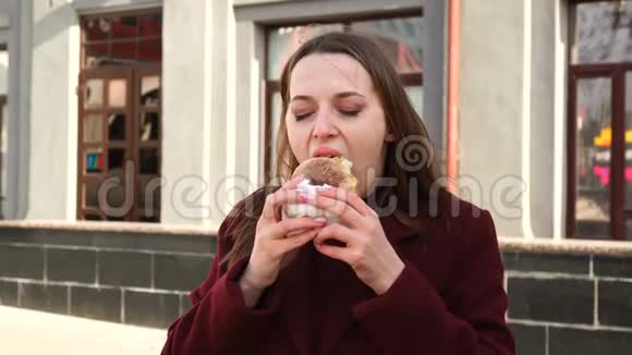 迷人的女孩在城市街道上愉快地吃着一个新鲜的汉堡视频的预览图