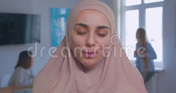 一位年轻美丽的微笑自信的穆斯林女孩在头巾办公室背景下的特写肖像视频的预览图