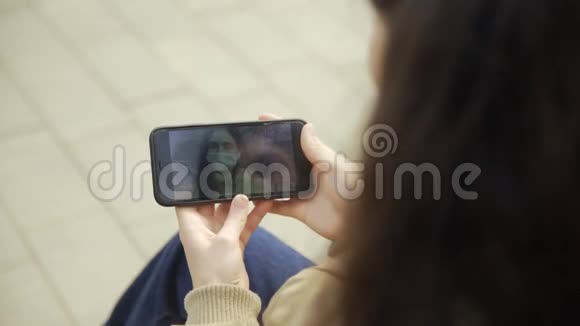 年轻女子戴着防护面具在城市公园里用智能手机观看视频大流行Covid19冠状病毒视频的预览图