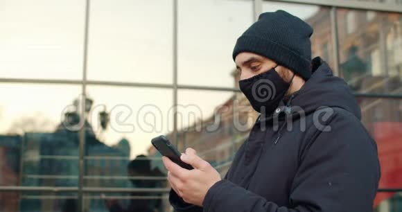 侧面观看男子戴黑色棉防护面具使用智能手机打字滚动和触摸手机屏幕视频的预览图
