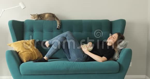 年轻女子躺在沙发上从玻璃碗里吃爆米花在家休息视频的预览图