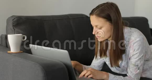 漂亮的年轻女人在沙发上喝咖啡或茶在家里的笔记本电脑上工作女孩在家工作自由职业者写作视频的预览图