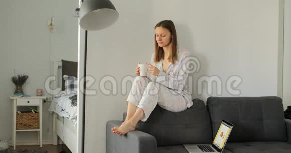 女孩在沙发上喝咖啡或茶女人在房间里工作学习和使用笔记本电脑自由职业写作视频的预览图