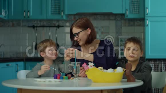 30岁的年轻母亲和两个儿子在家里的蓝色厨房里画复活节彩蛋准备复活节的幸福家庭视频的预览图