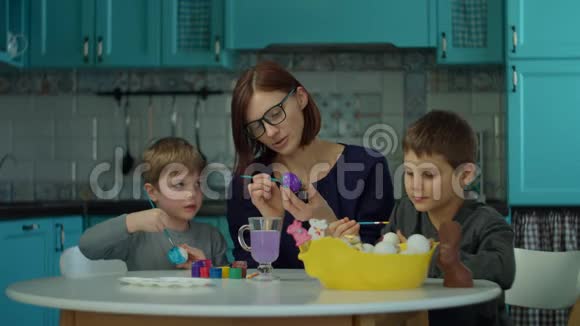 30岁的年轻母亲和两个儿子在家里的蓝色厨房里画复活节彩蛋准备复活节的幸福家庭视频的预览图